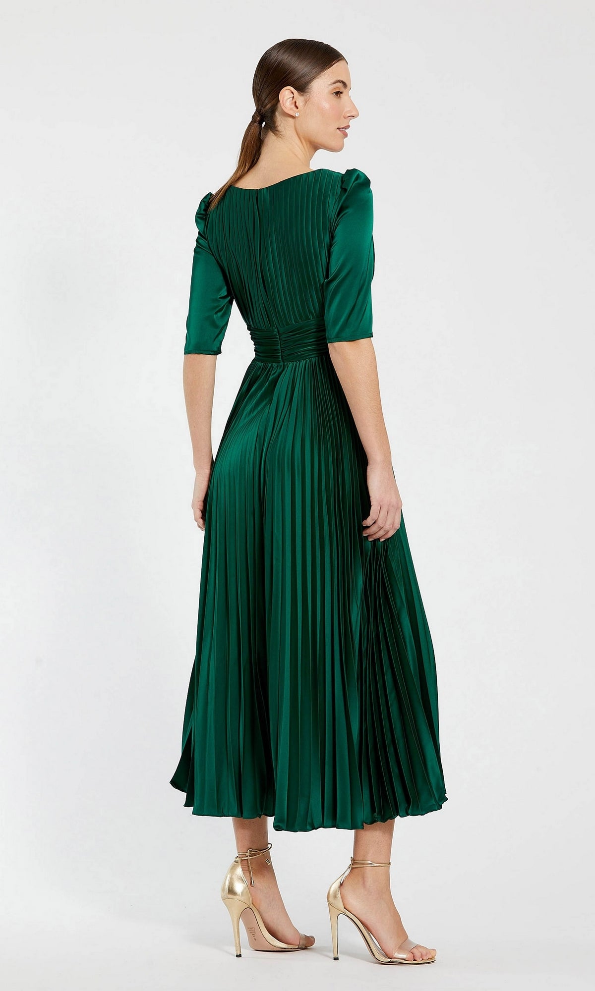 A woman stands facing away, wearing the Long Formal Dress 27341 by Mac Duggal in emerald green—a pleated a-line midi dress with elbow-length sleeves—paired with gold high-heeled sandals.